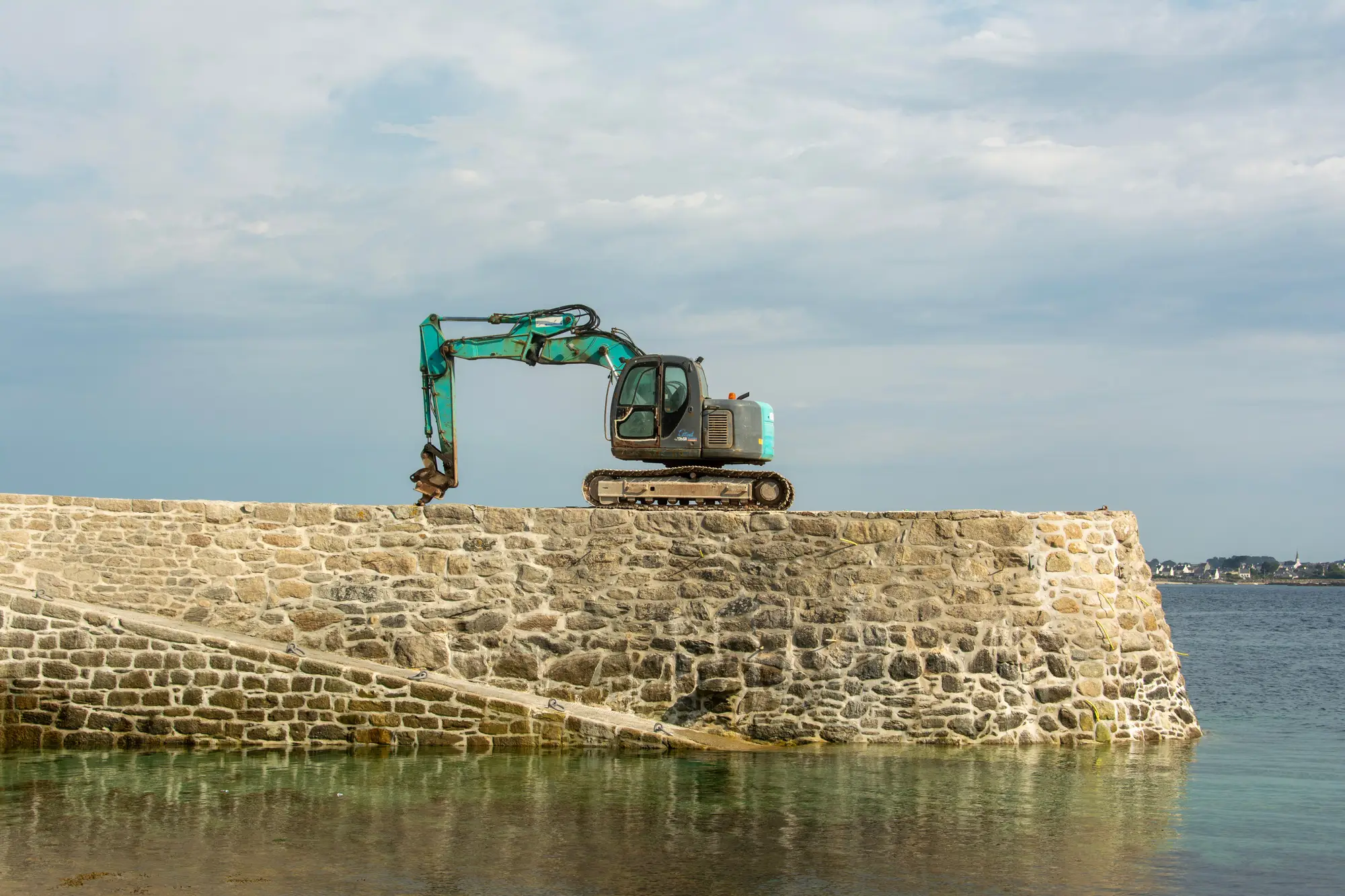 An excavator parked on top of a stone quay wall, with shallow water below